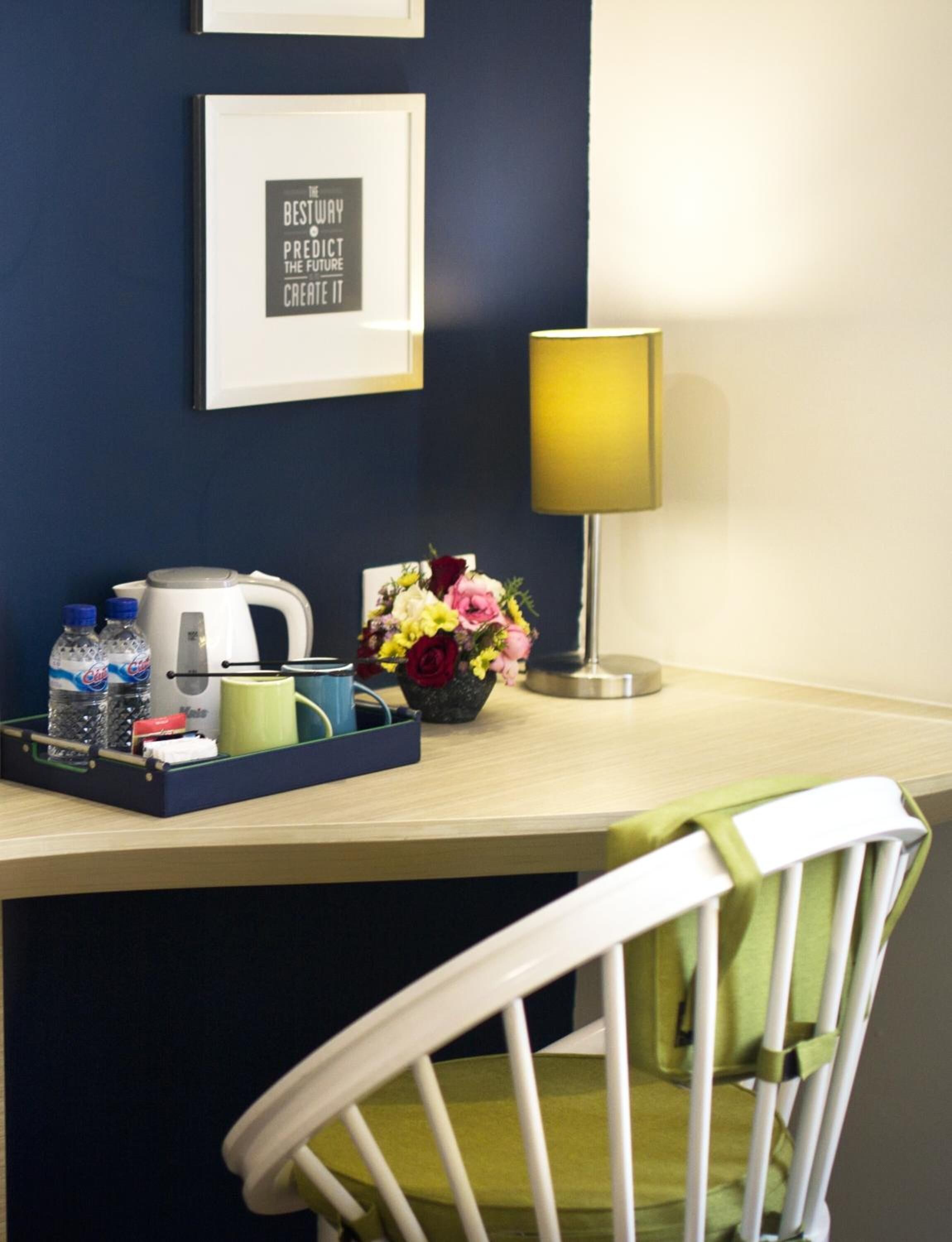 A hotel room with a desk, a framed inspirational quote, a lamp, a tray with toiletries, and a flower vase.