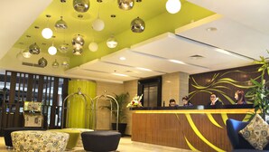 Zona de recepción de hotel con techo verde, iluminación colgante moderna, mostrador de recepción y asientos cómodos.