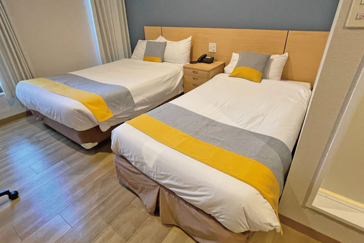 A hotel room with two beds, each with a wooden headboard, a bedside table with a phone, and a light-colored wooden floor.