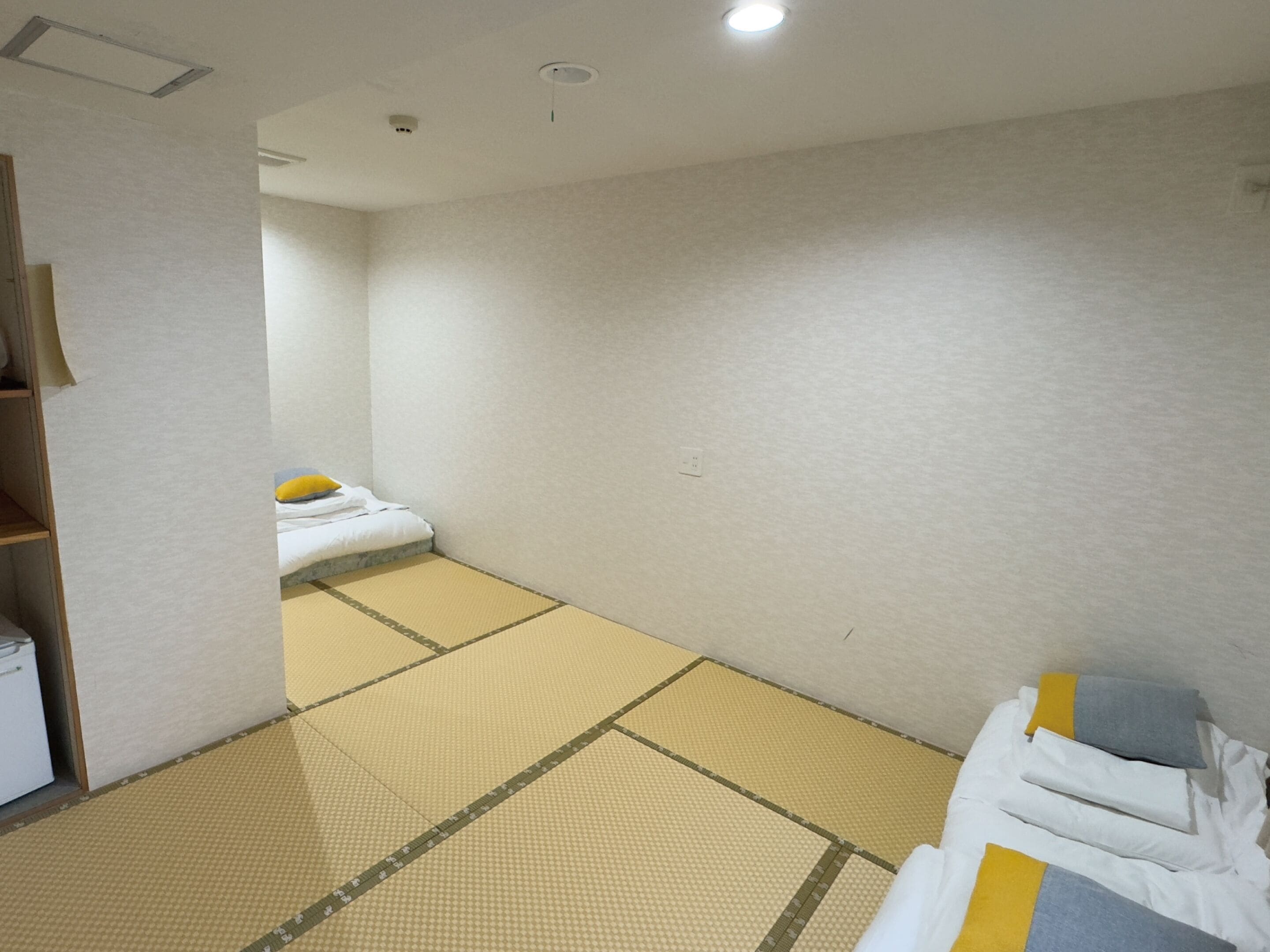 Une chambre d’hôtel avec un sol recouvert de tatamis, un lit avec une literie blanche et un oreiller jaune, et une petite armoire en bois.