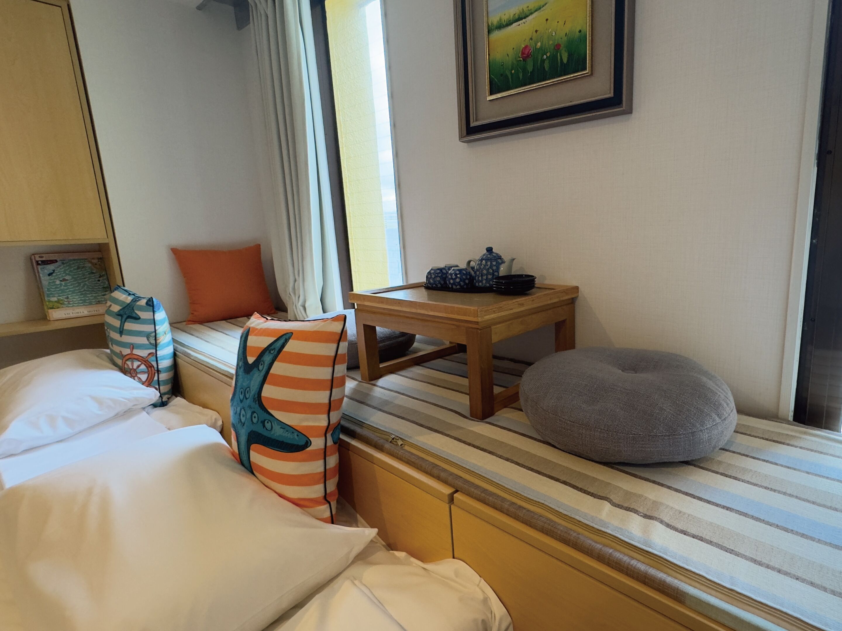 A hotel room with a bed, a wooden nightstand, a gray bean bag, a window with curtains, and a framed picture on the wall.