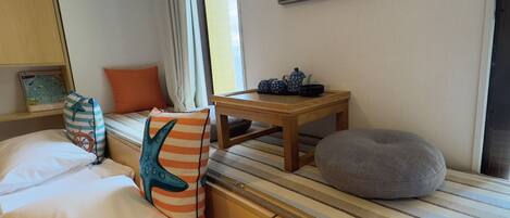 A hotel room with a bed, a wooden nightstand, a gray bean bag, a window with curtains, and a framed picture on the wall.