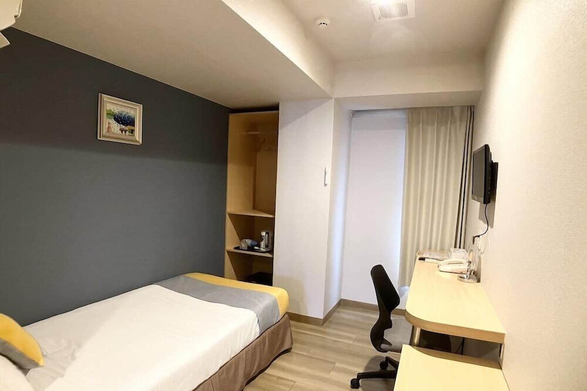 A compact hotel room with a single bed, a desk, a chair, a television, and a small shelf with personal items.