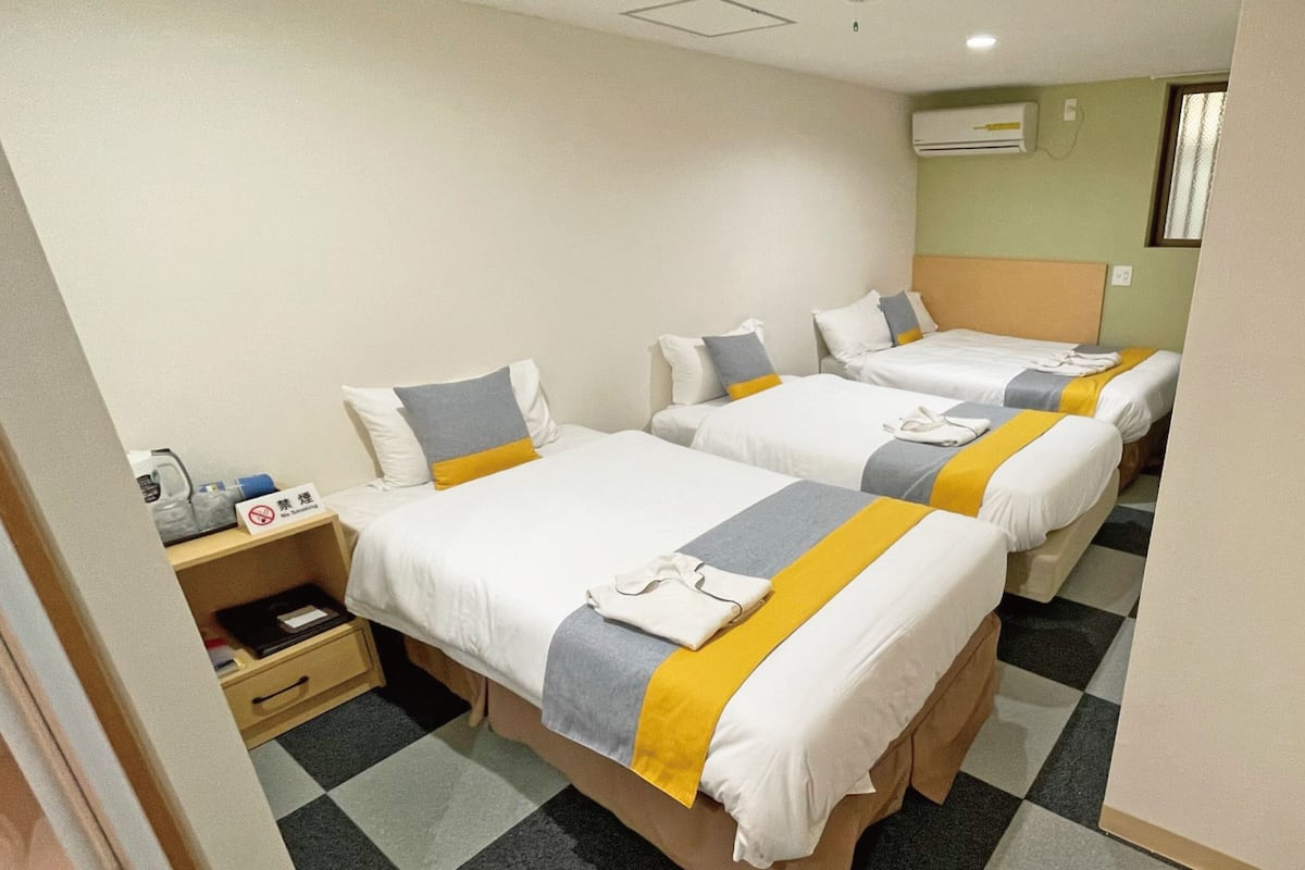 A hotel room with three single beds, a small desk with a chair, and a wall-mounted air conditioner.