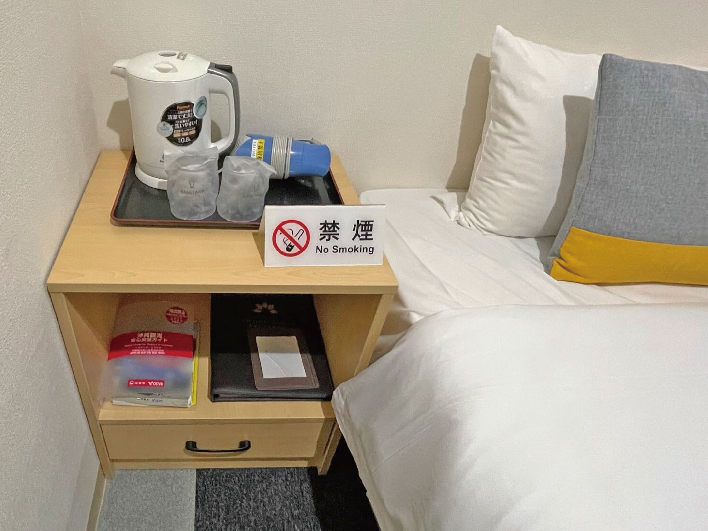 A hotel room with a bed, a nightstand with a coffee maker, a tray with a water bottle, and a 'No Smoking' sign.