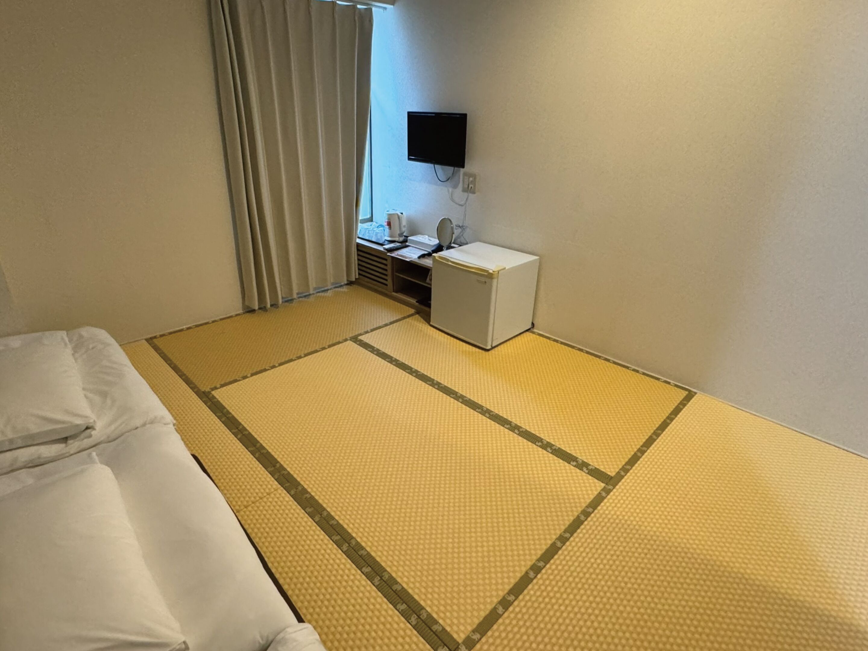 A hotel room with tatami mat flooring, a bed, a small desk with a mirror, and a television mounted on the wall.