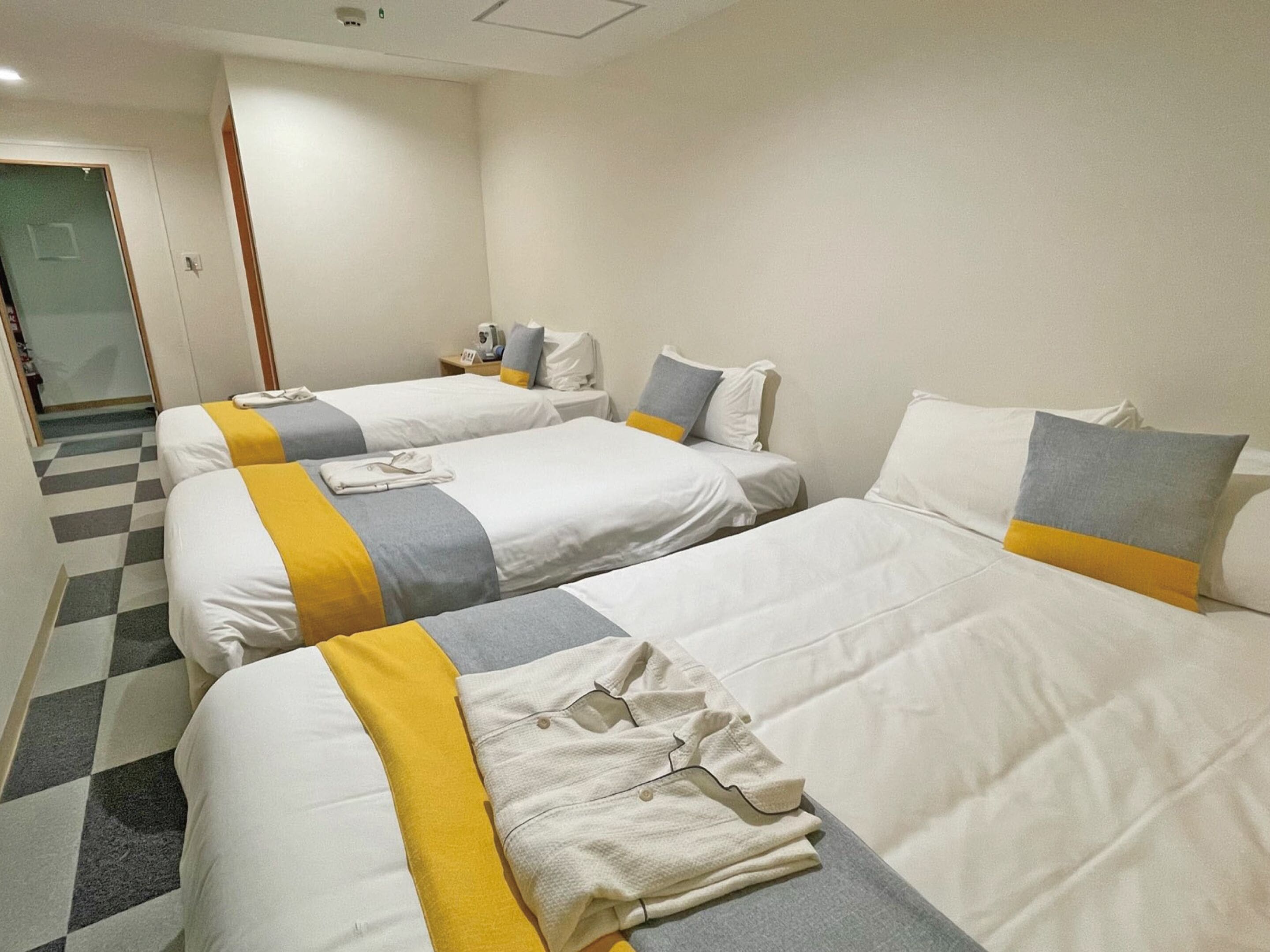 A hotel room with two beds, each with white bedding and a yellow accent. There is a small table with a phone and a lamp.