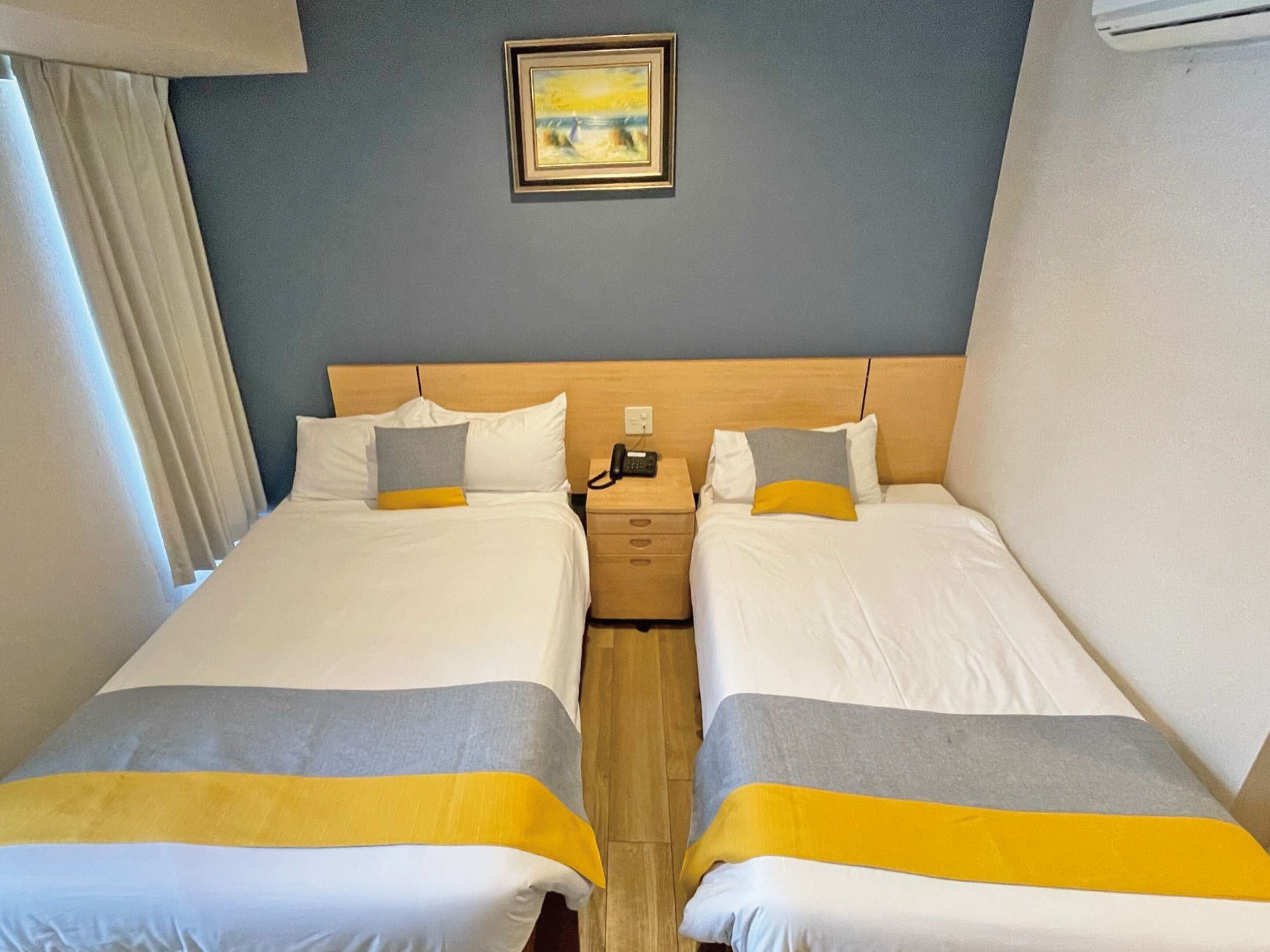 Quarto de hotel com duas camas, cada uma com roupa de cama branca e detalhes amarelos. Há uma pintura na parede acima das camas, uma mesa de cabeceira com telefone e uma janela com cortinas.