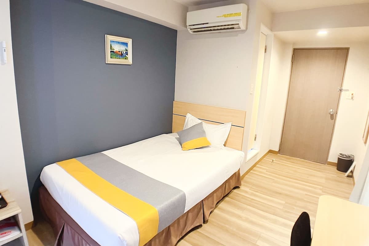 A hotel room with a bed, a desk, a chair, an air conditioning unit, and a door.
