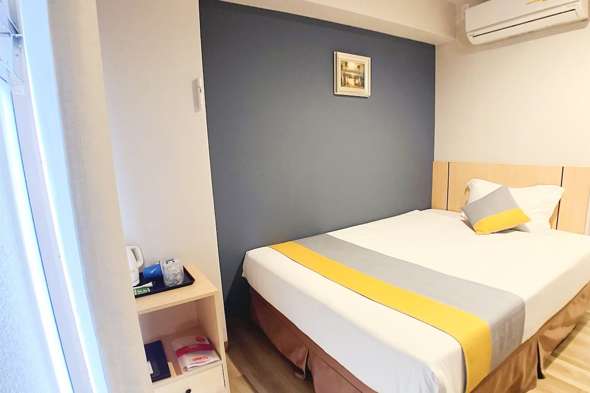 A hotel room with a bed, a bedside table, a small shelf with items, a wall-mounted air conditioner, and a picture on the wall.