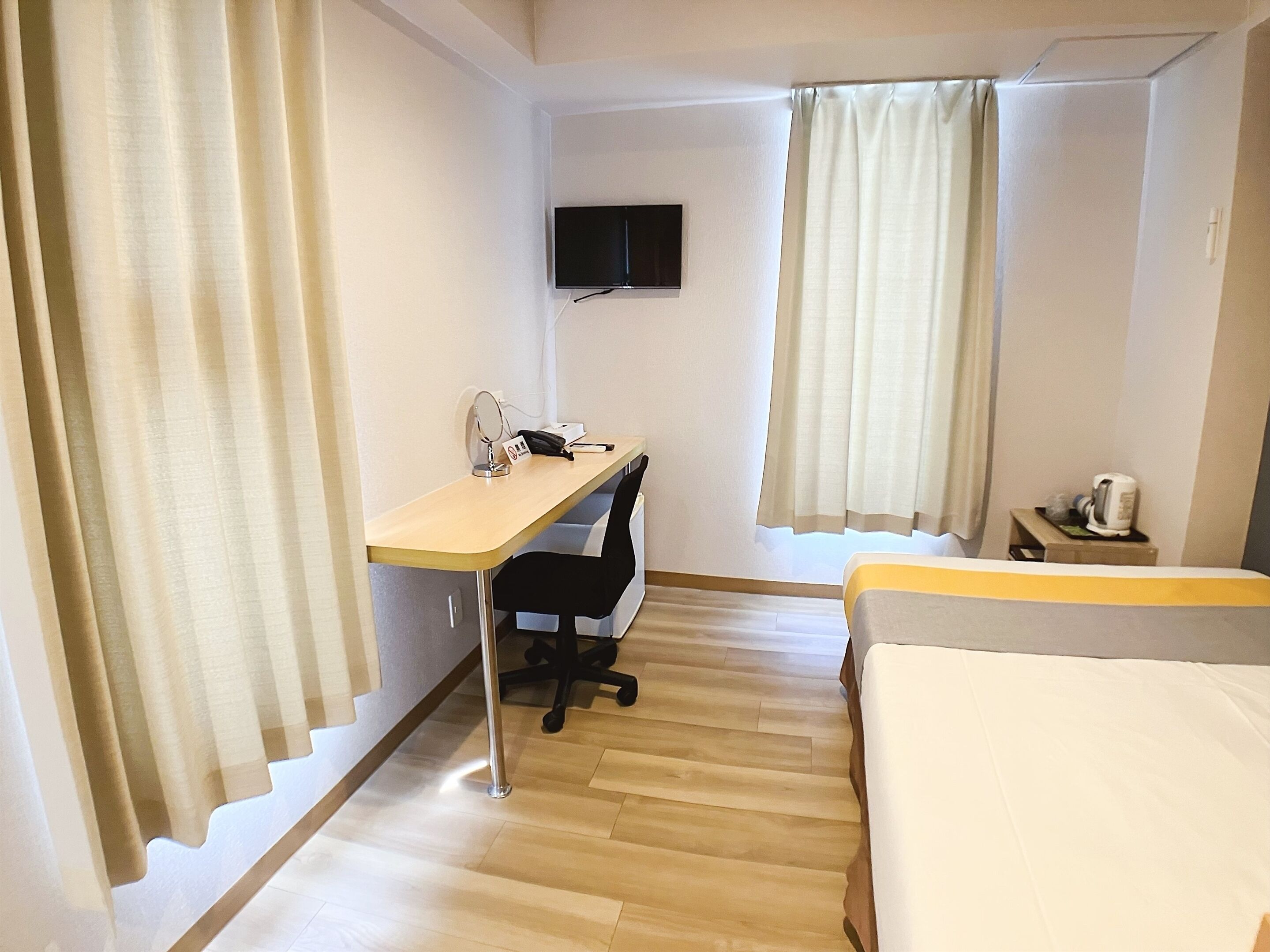 Una habitación de hotel con un escritorio de madera, una silla, una cama, un televisor montado en la pared y una ventana con cortinas.