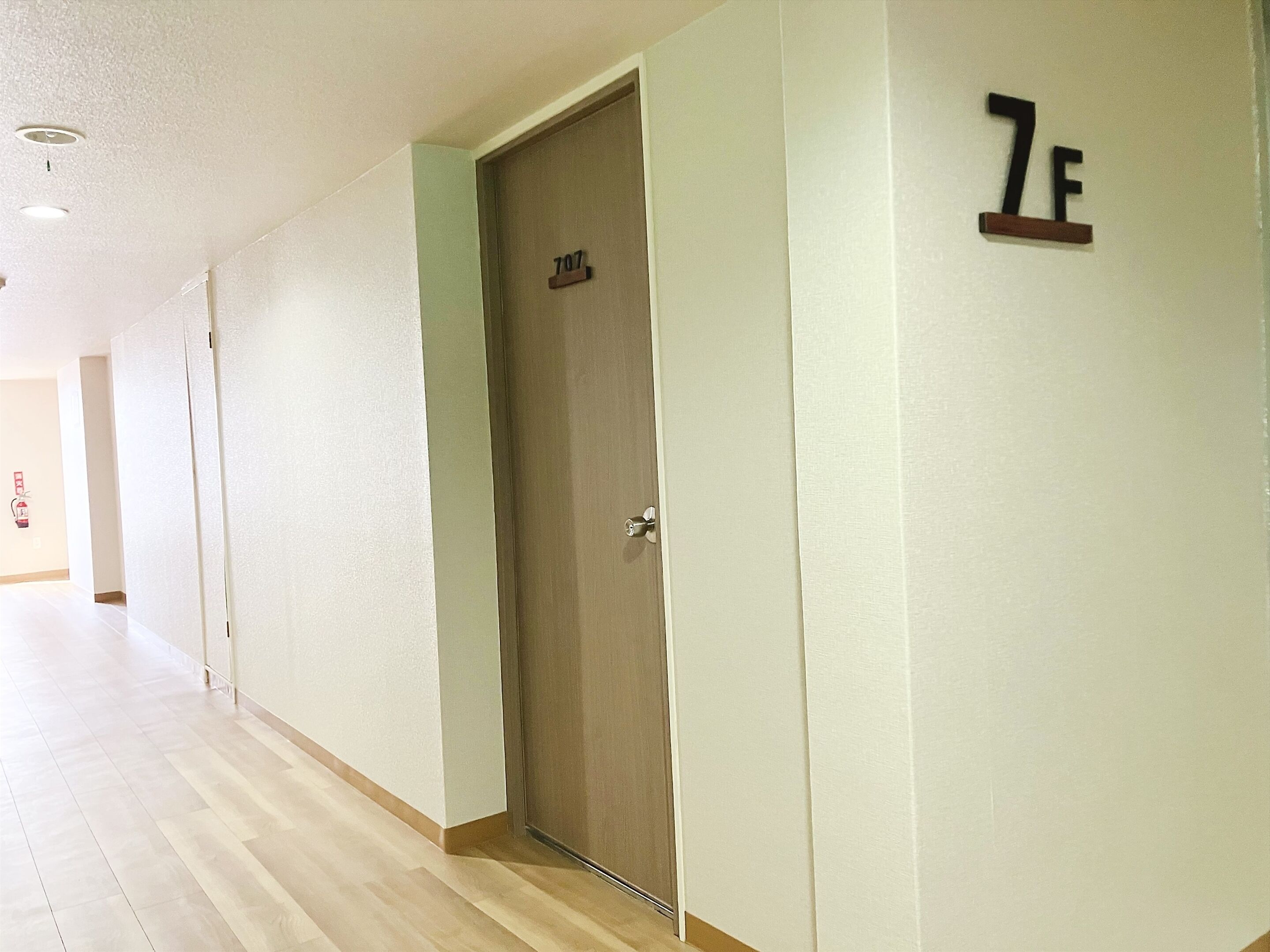Een gang met een houten deur waarop het nummer 707 staat, witte muren en een lichtkleurige houten vloer.
