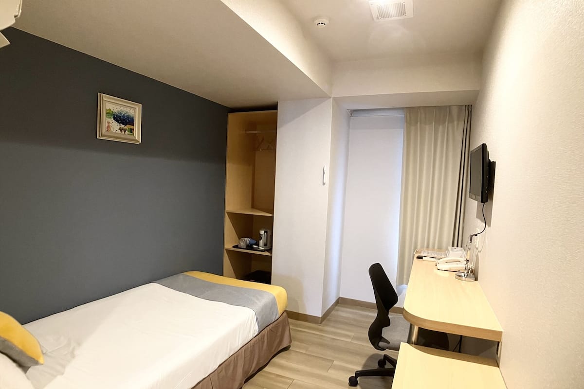 A small, neatly arranged bedroom with a bed, a desk, and a shelf unit.