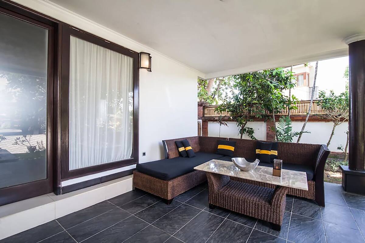 A covered outdoor seating area with a sofa and a table, a large window with curtains, and a view of greenery and buildings in the background.