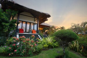 A traditional house with a thatched roof, a balcony with red chairs, and a lush garden with various plants and flowers. - Villa L'Orange Bali (Gianyar)
