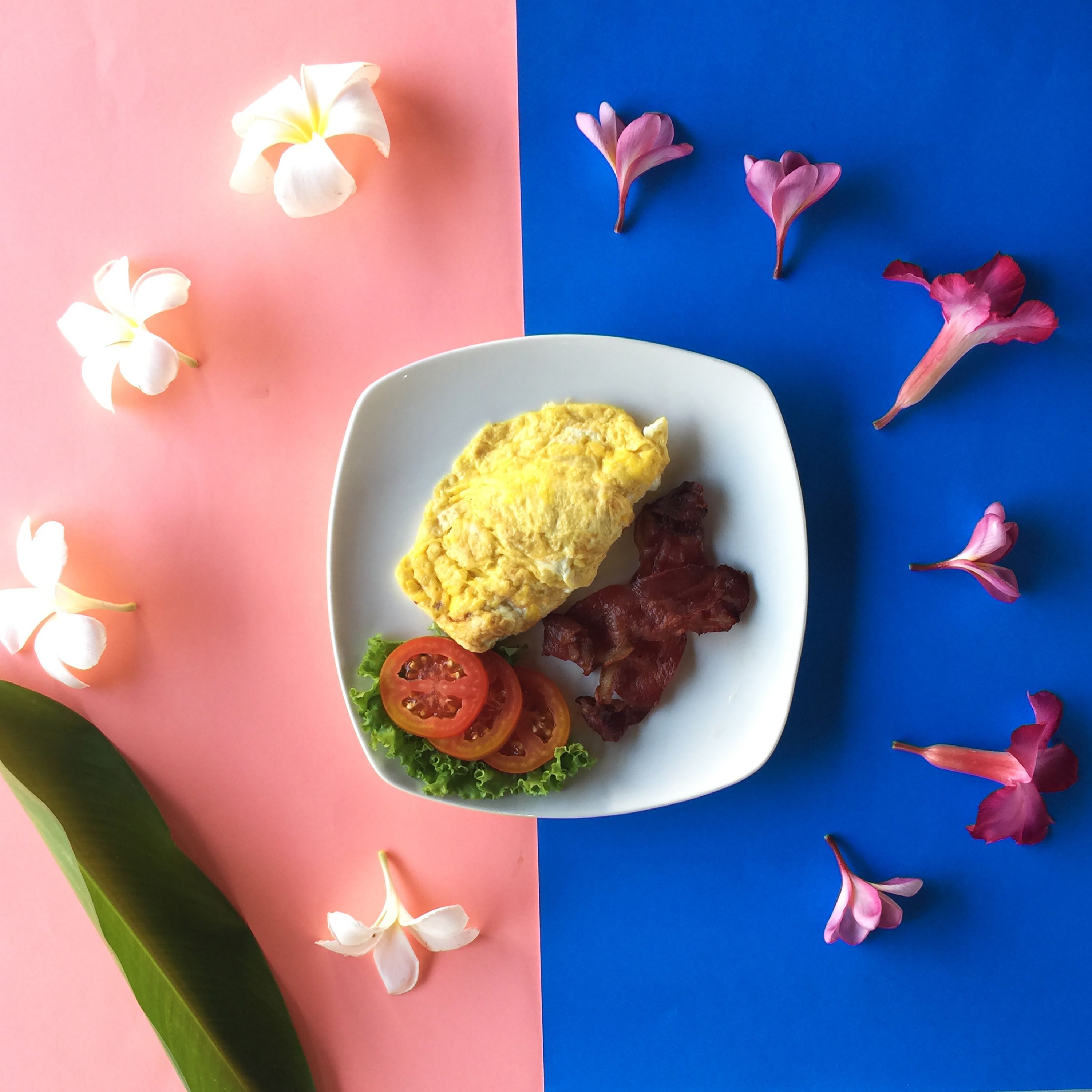 Prato com ovos mexidos, bacon e rodelas de tomate em um prato branco, rodeado por flores sobre um fundo rosa e azul.