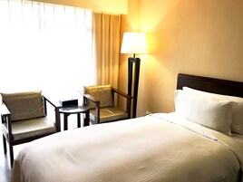 Una habitación de hotel con cama, dos sillones, una mesita, una lámpara y una ventana con cortinas.