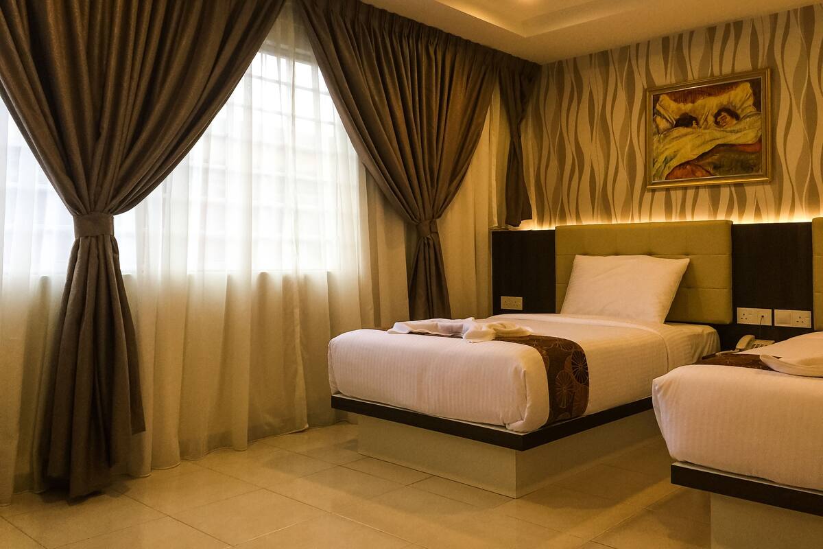 Habitación de hotel con dos camas, un ventanal grande con cortinas, un cuadro en la pared y una mesita de noche con un teléfono.