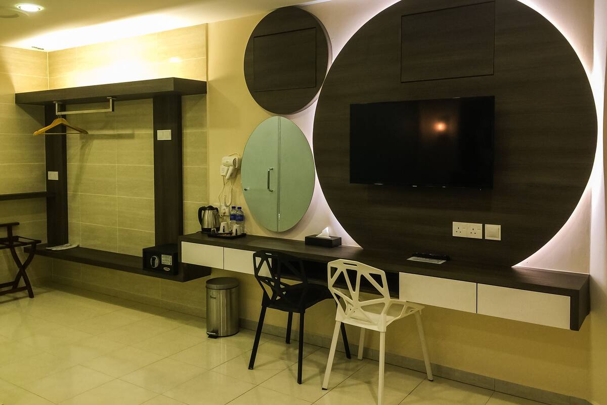 Una habitación de hotel moderna con un espejo grande, un televisor de pantalla plana, un escritorio con silla, un perchero y un cesto de basura.
