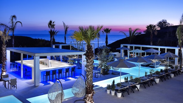 Un moderno espacio exterior con bar, asientos y piscina, con vistas al océano al atardecer.