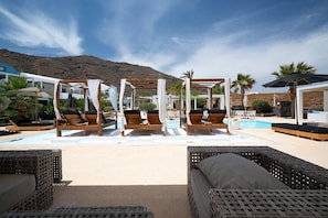 Zona de piscina con tumbonas de madera, un toldo y vistas a un paisaje montañoso.