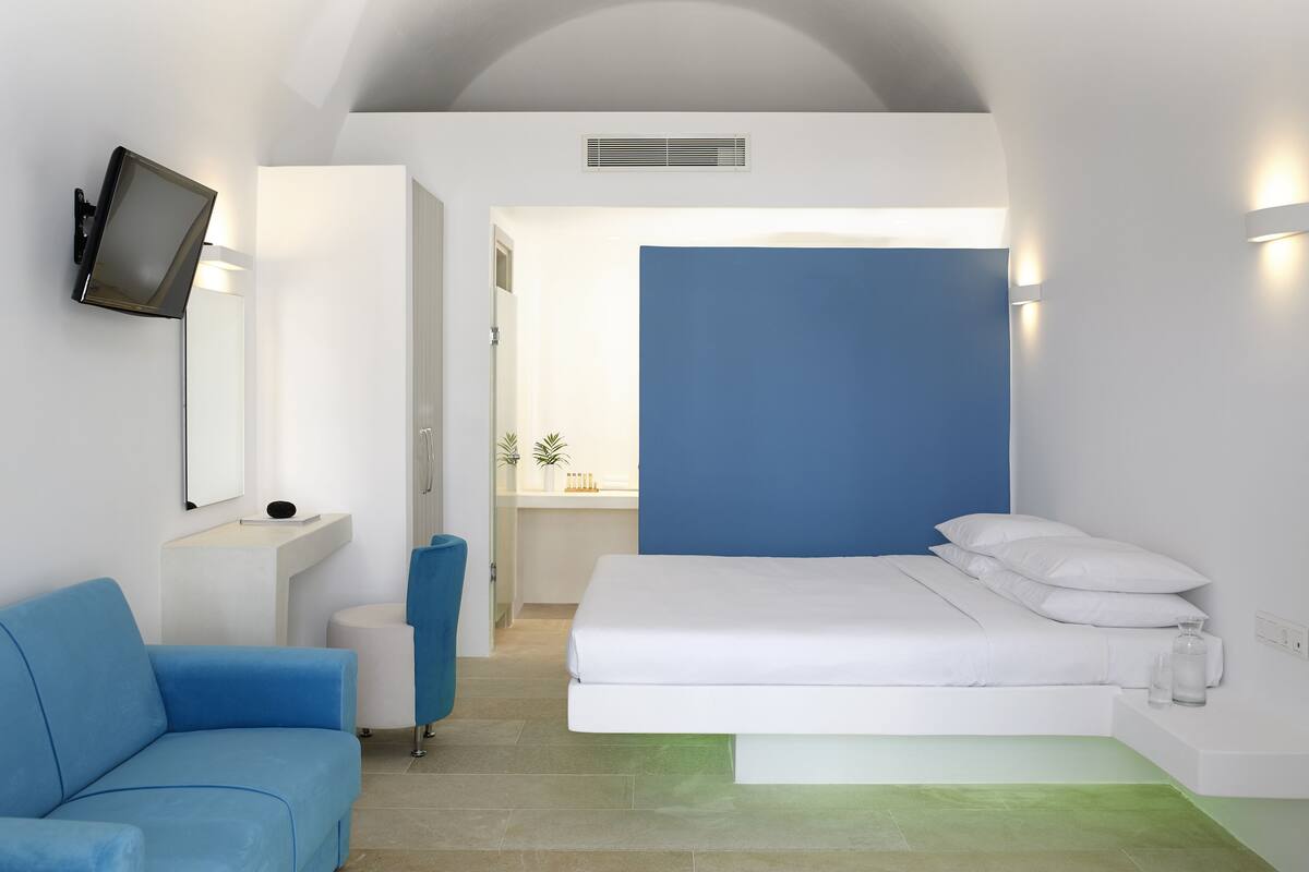 Une chambre d’hôtel moderne avec un lit, un canapé bleu, un téléviseur fixé au mur et une salle de bain visible à travers une porte ouverte.