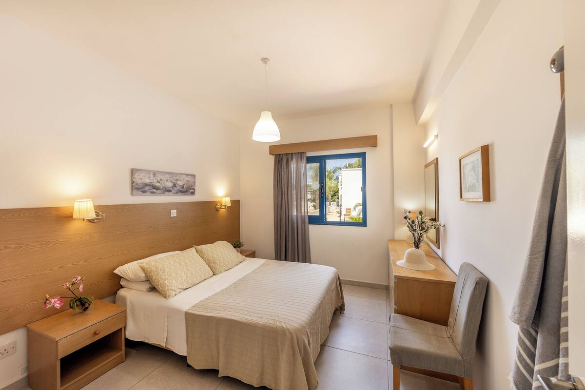 A bedroom with a bed, bedside tables, a nightstand, a desk, a chair, a window with curtains, and a hanging light fixture.