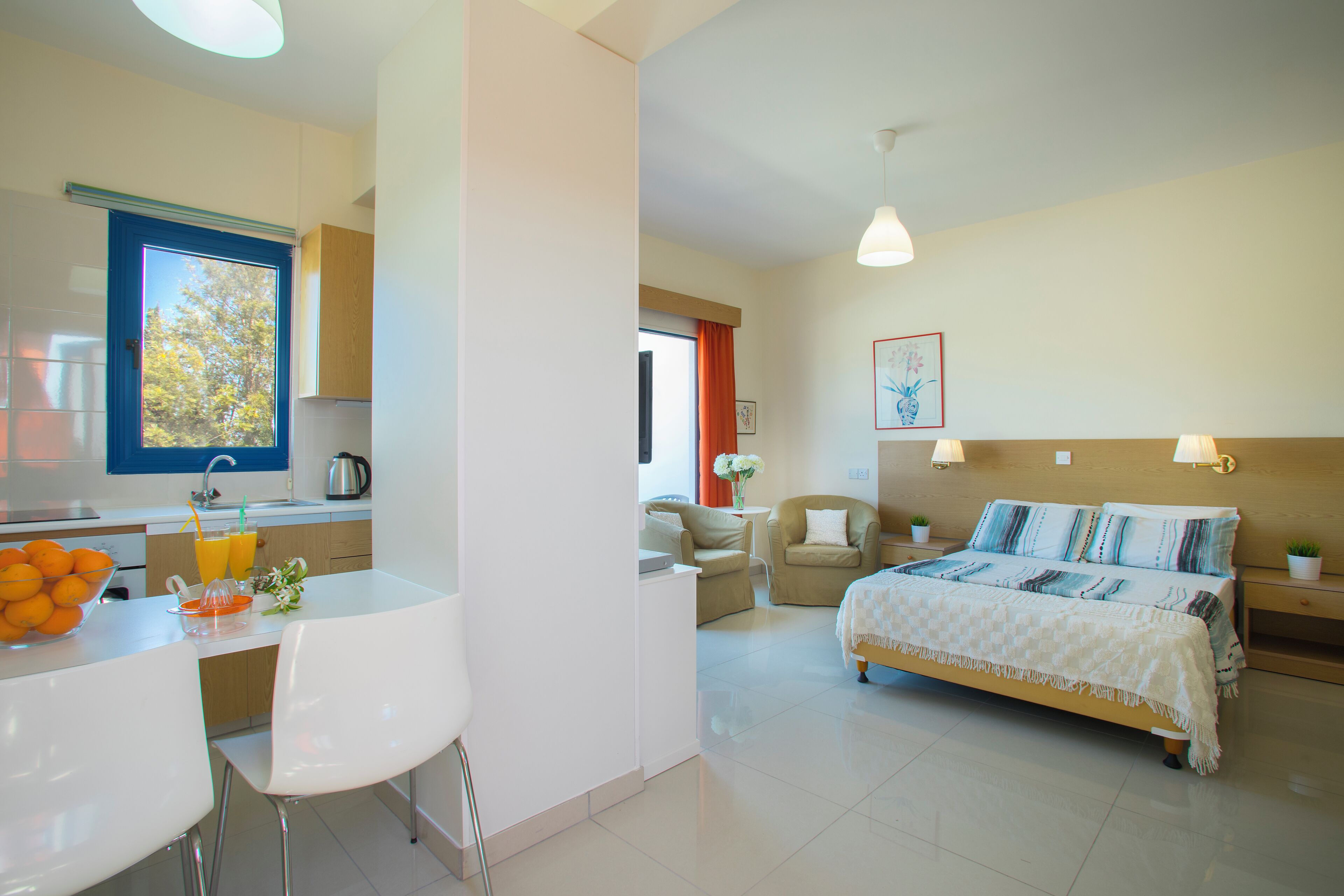A modern hotel room with a kitchen area, a dining table with chairs, a bed with a headboard, and a small table with a vase of flowers.