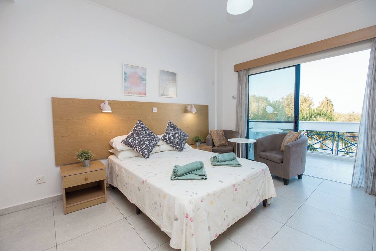 A bedroom with a bed, bedside tables, a small table, a chair, and a balcony with a view.