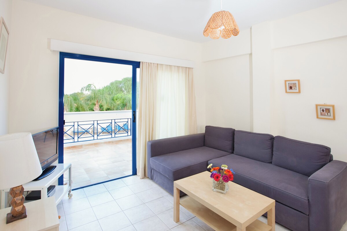 A modern living room with a grey sofa, a wooden coffee table with a vase of flowers, a TV on a white stand, and a balcony with a blue railing.