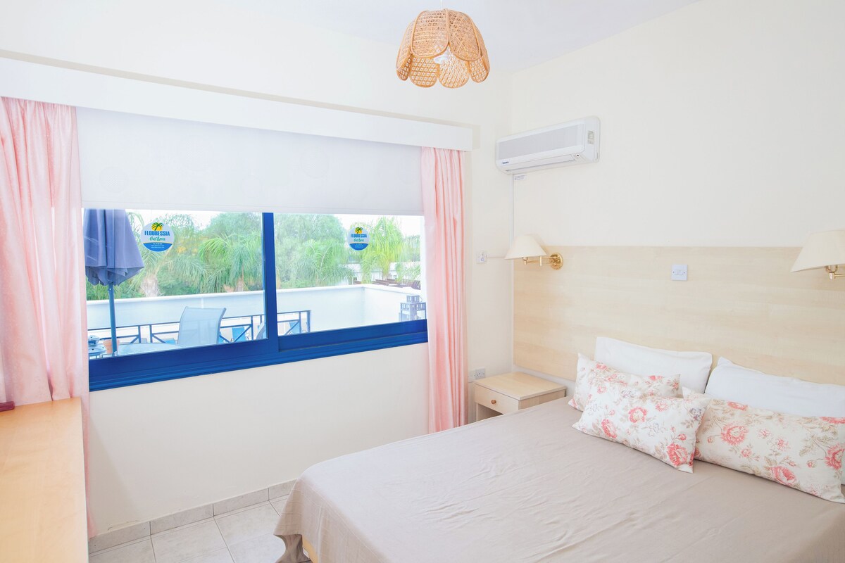 A hotel room with a bed, a nightstand, a lamp, a window with curtains, and a view of a pool and greenery outside.