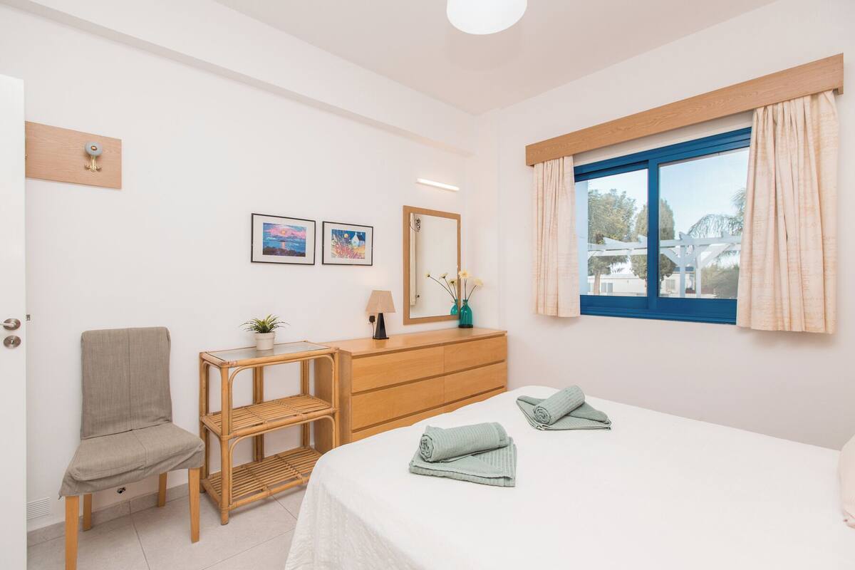 A bedroom with a bed, a chair, a small table, a dresser, and a window with curtains.