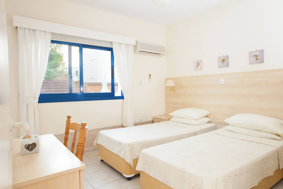 A hotel room with two single beds, a wooden chair, a small table, and a window with curtains.