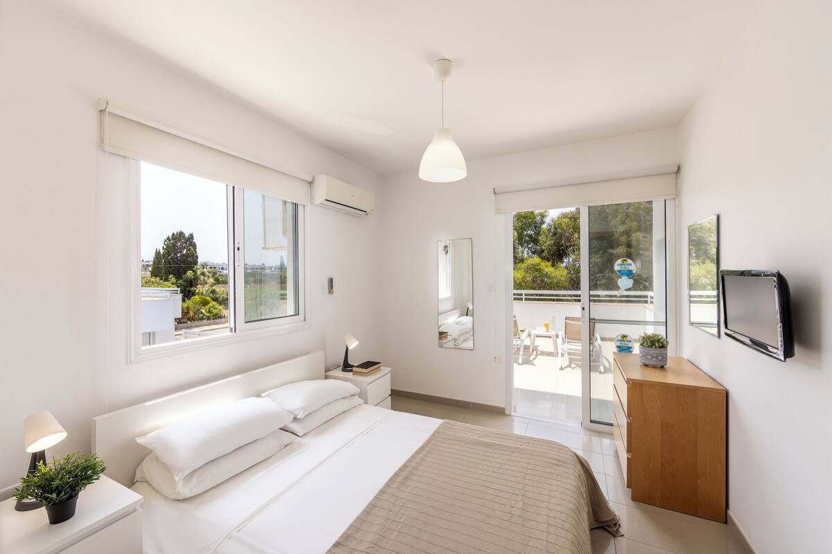 A modern bedroom with a bed, bedside table, lamp, and a view of the outdoors through a sliding glass door.