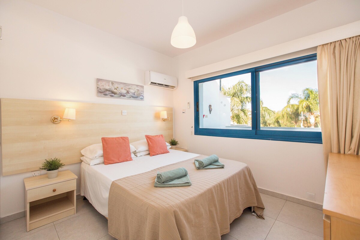 A bedroom with a bed, bedside tables, a window with a view, and an air conditioning unit.