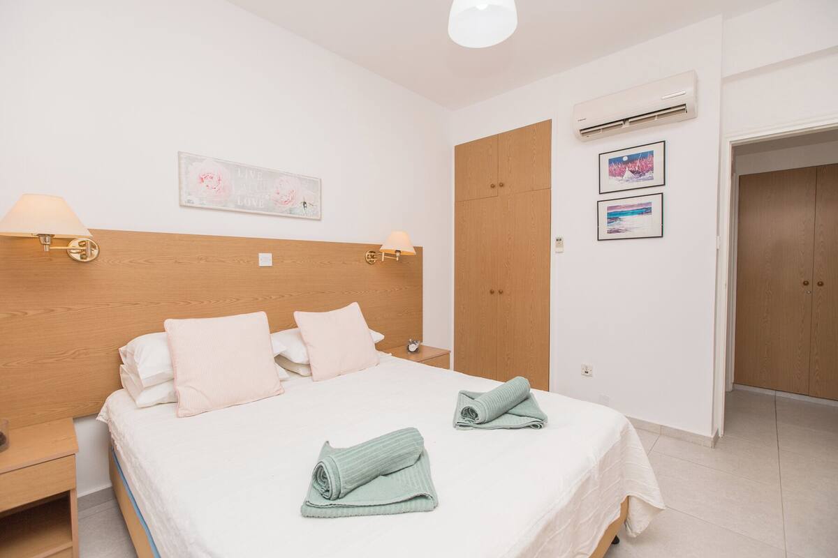 A neatly made bed with pillows and towels, a wooden headboard, bedside lamps, and a wall-mounted air conditioner.