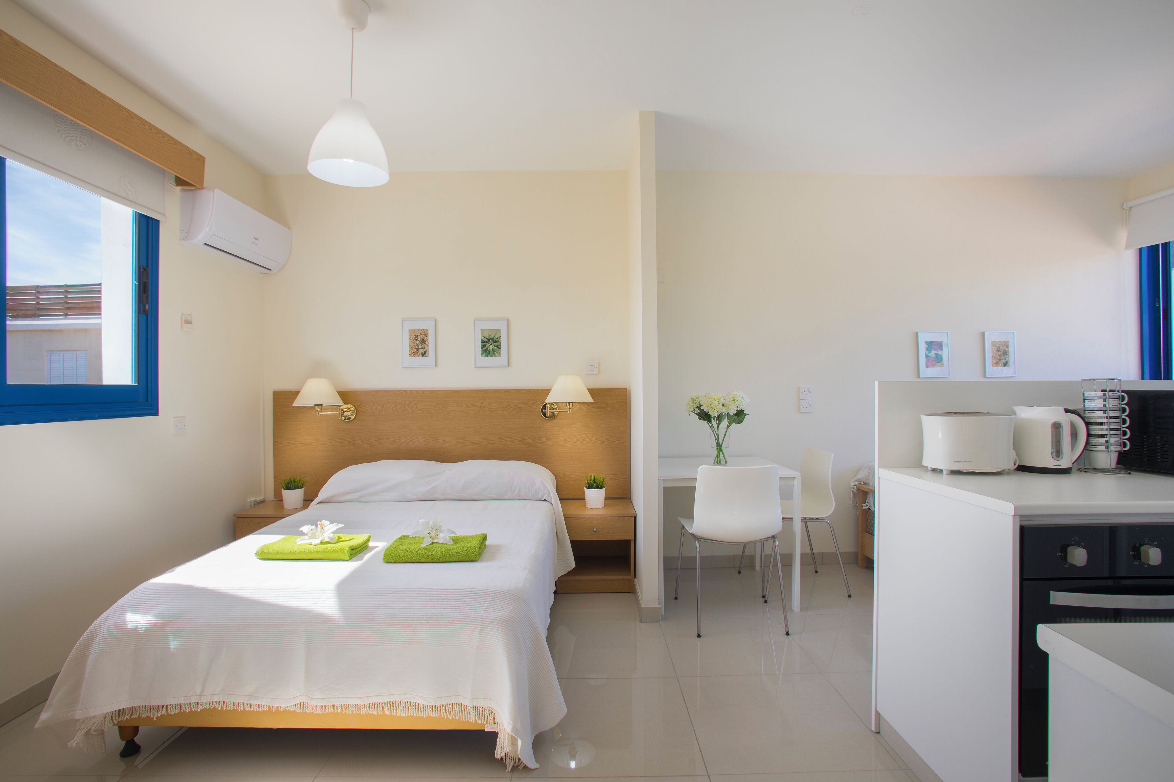 Quarto de hotel com cama, escrivaninha, cadeira, cozinha compacta e janela.