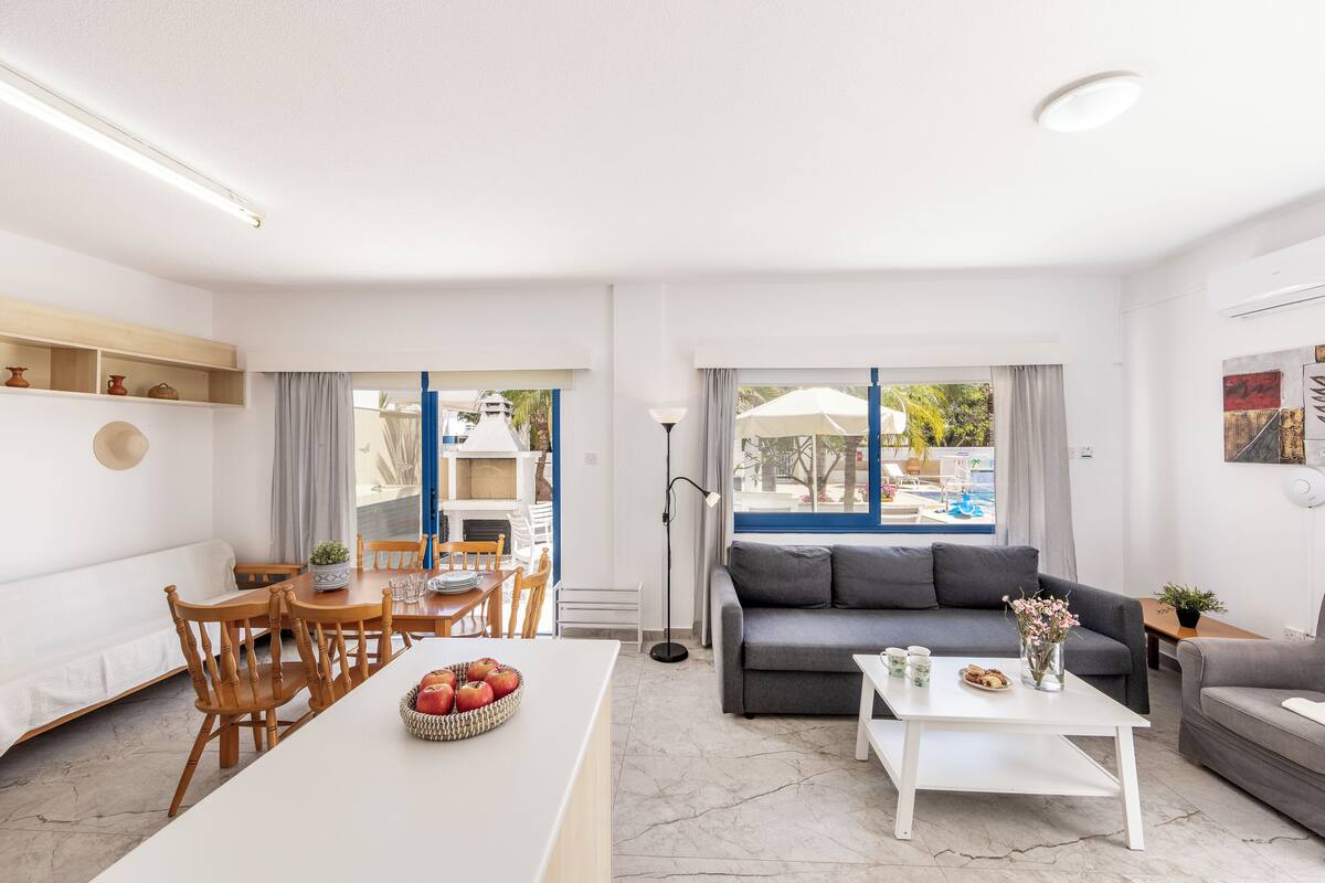 A modern living room with a sofa, dining table, and chairs, a kitchen area, and large windows with a view of the outdoors.