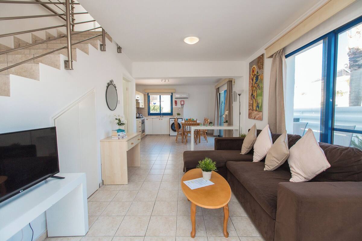 A modern living room with a TV, a sofa, a coffee table, and a dining area with a kitchen in the background.