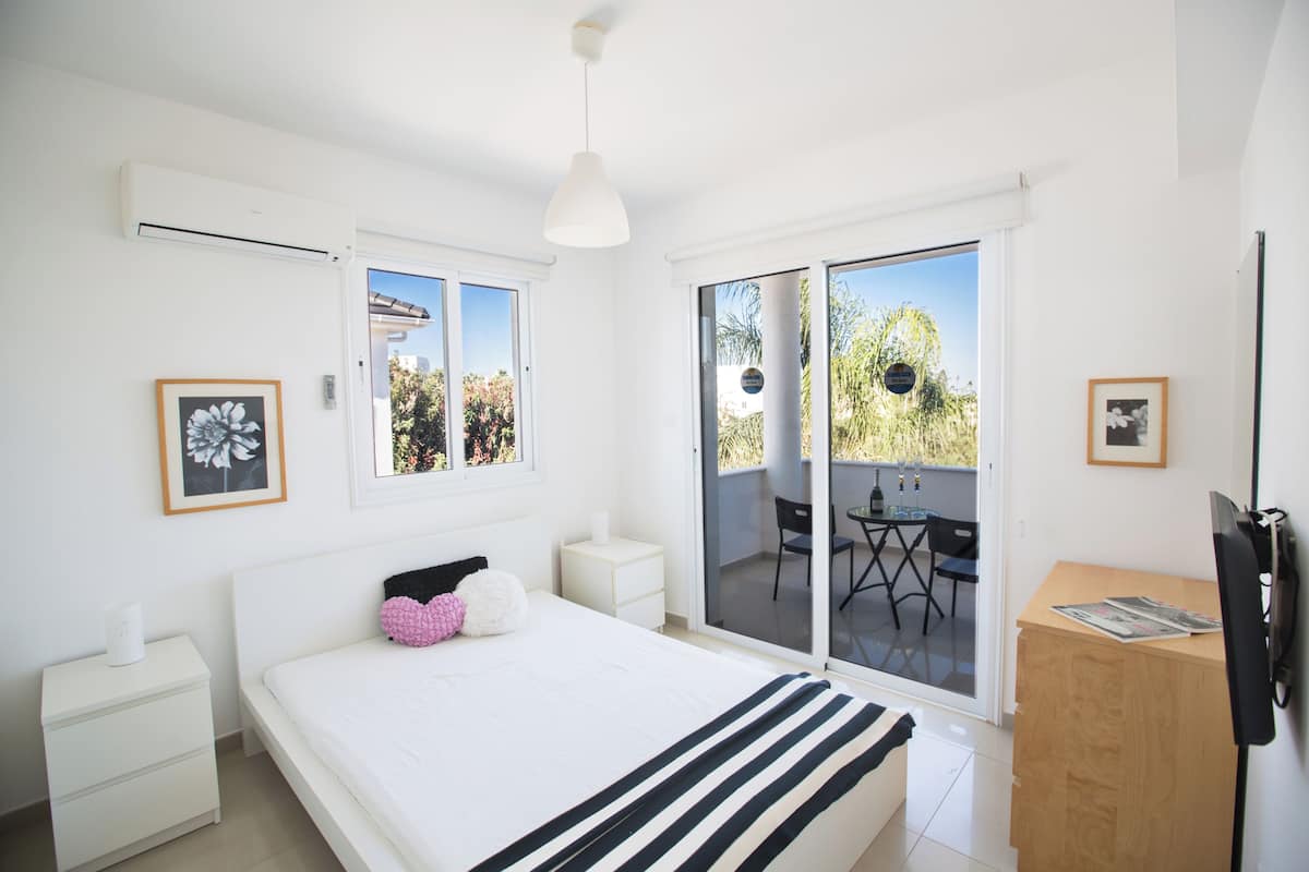 A bedroom with a bed, bedside tables, a desk, and a sliding glass door leading to an outdoor area with a table and chairs.