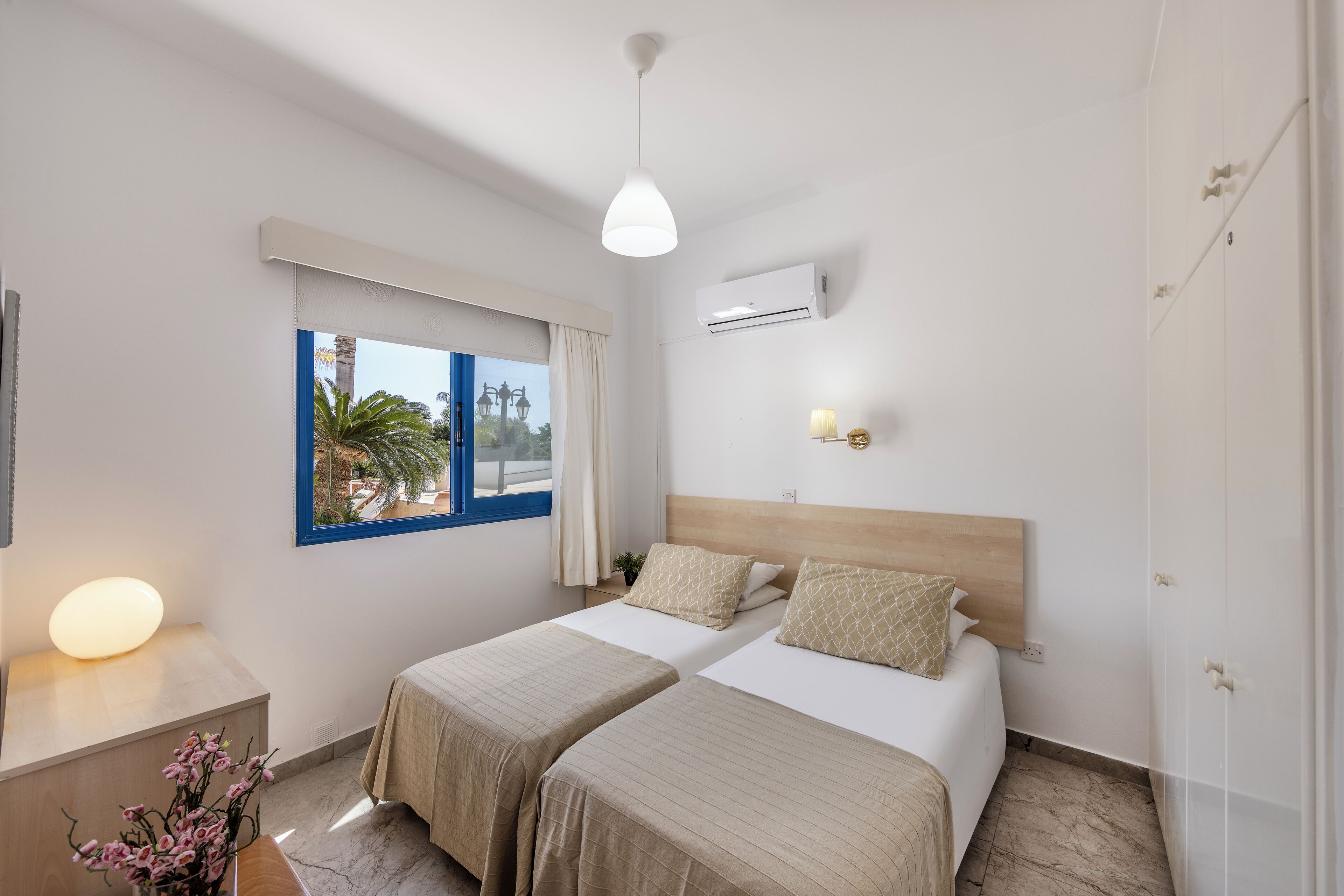 A hotel room with two beds, a window with a view, an air conditioning unit, and a bedside lamp.