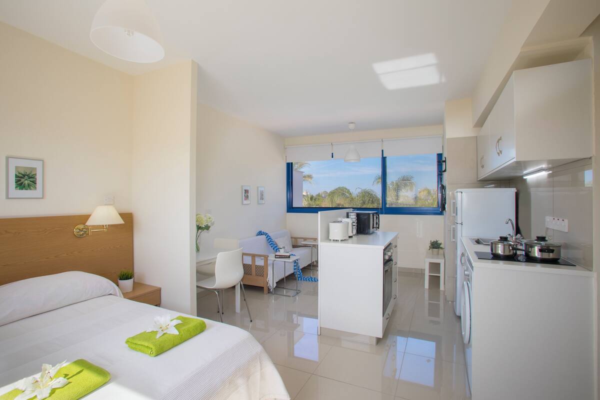A hotel room with a bed, a desk, a kitchenette, and a view of palm trees.
