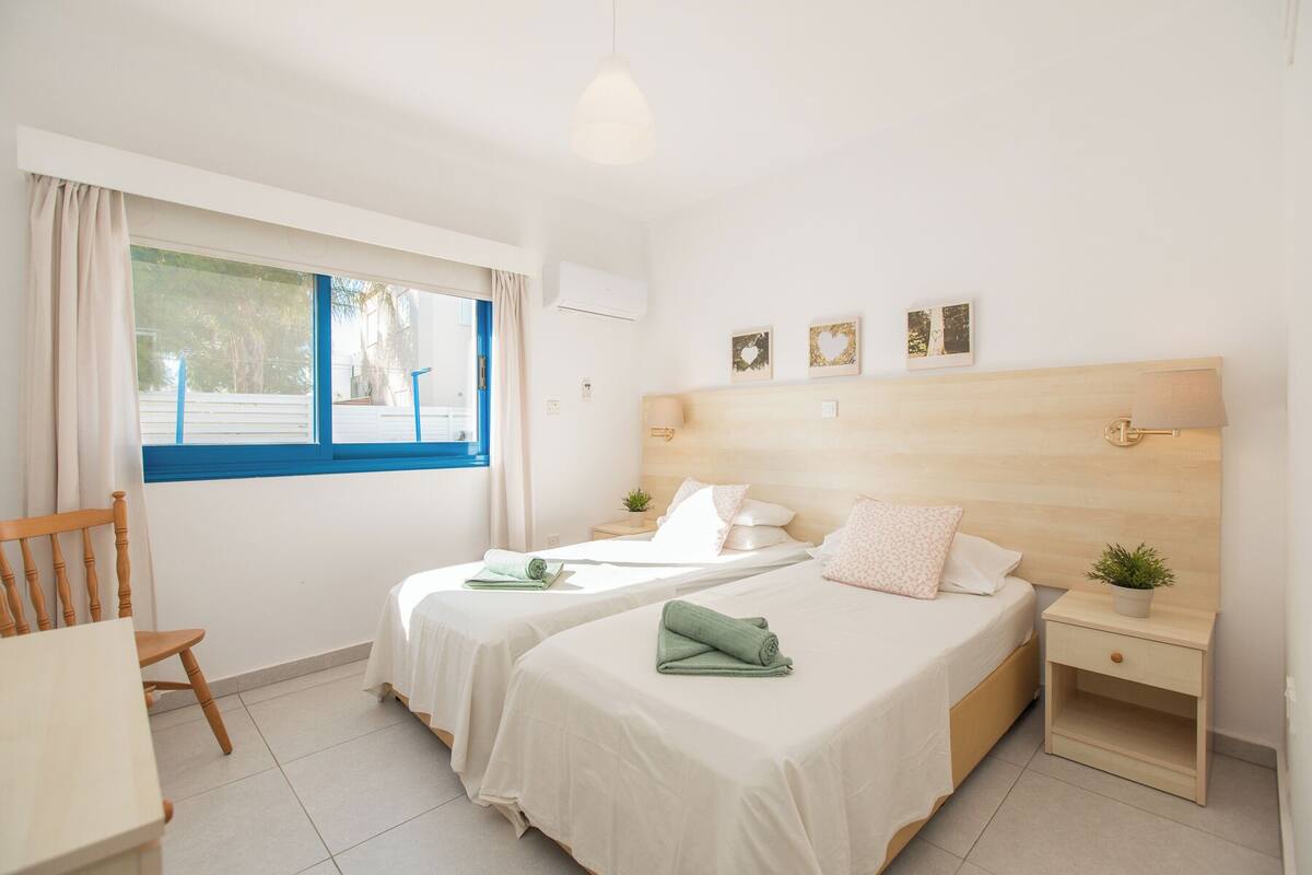 A hotel room with two beds, a small table, a chair, a bedside table with a lamp, and a window with curtains.