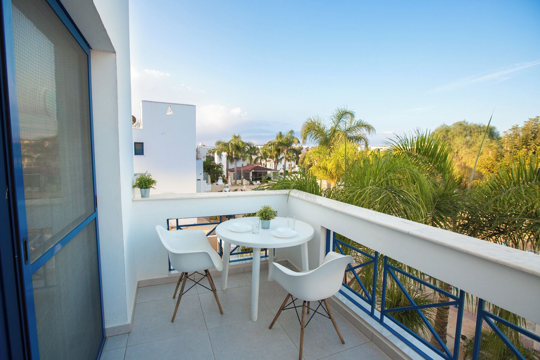 A balcony with a white table and two white chairs, a blue railing, and a view of palm trees and buildings.