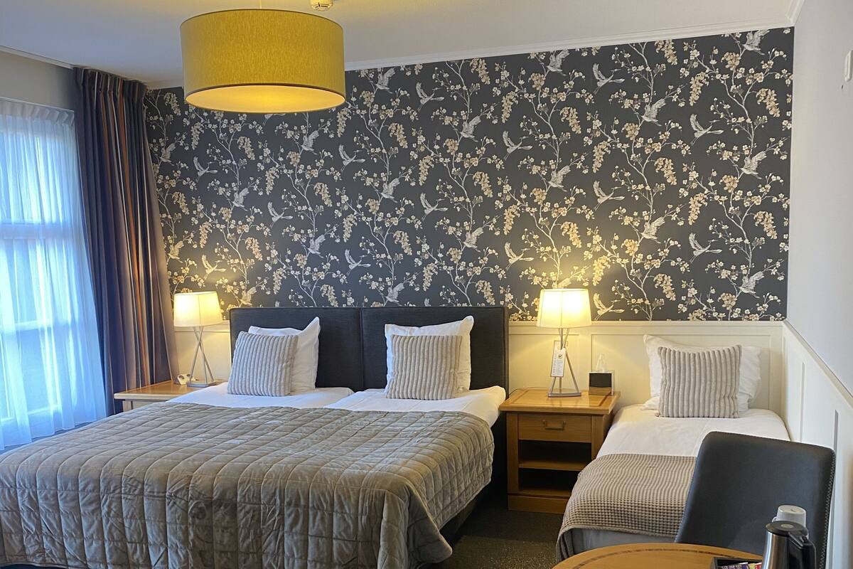 Une chambre d’hôtel avec deux lits, une table ronde en bois et une chaise. Il y a deux tables de chevet avec des lampes.