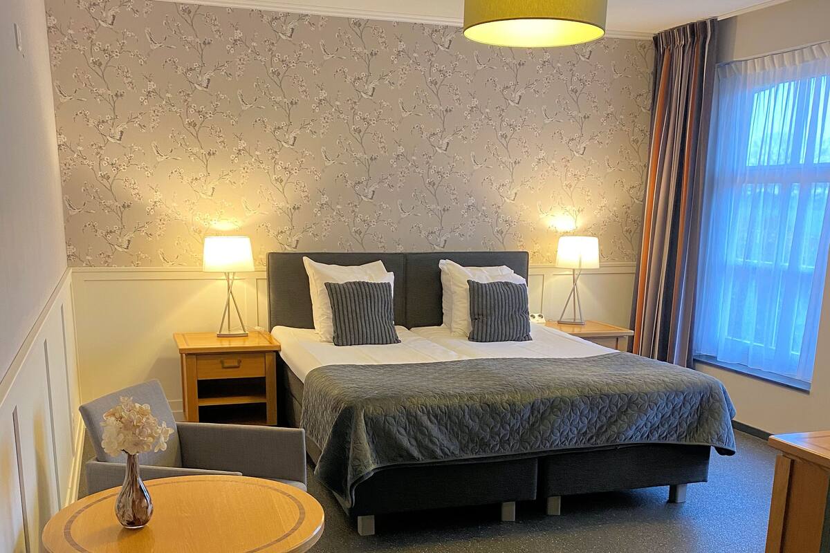 Une chambre d’hôtel comprenant un lit, deux lampes de chevet, une table de chevet, une chaise, une petite table avec un vase et une fenêtre avec des rideaux.