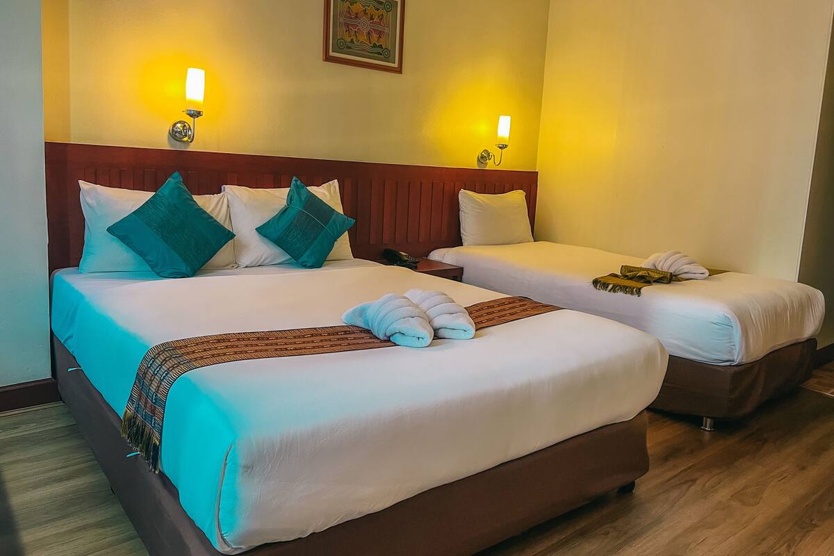 A hotel room with two beds, each with a wooden headboard, white linens, and teal pillows. There are two wall-mounted lamps and a framed artwork above the beds.