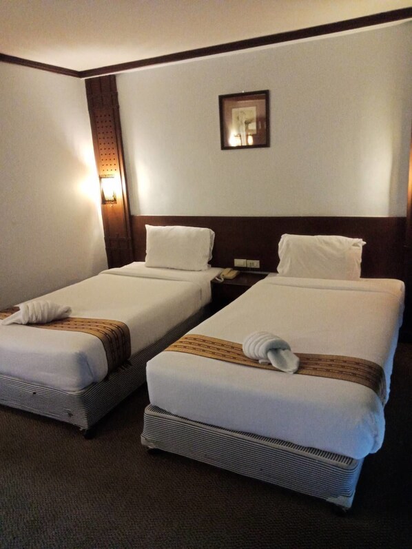 A hotel room with two beds, each with a white bedspread and a brown decorative strip. There is a nightstand with a lamp between the beds, and a framed picture on the wall above the beds.