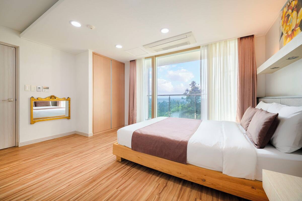 A modern bedroom with a wooden floor, a large bed with a brown blanket, a bedside table, a mirror, and a window with a view of greenery.
