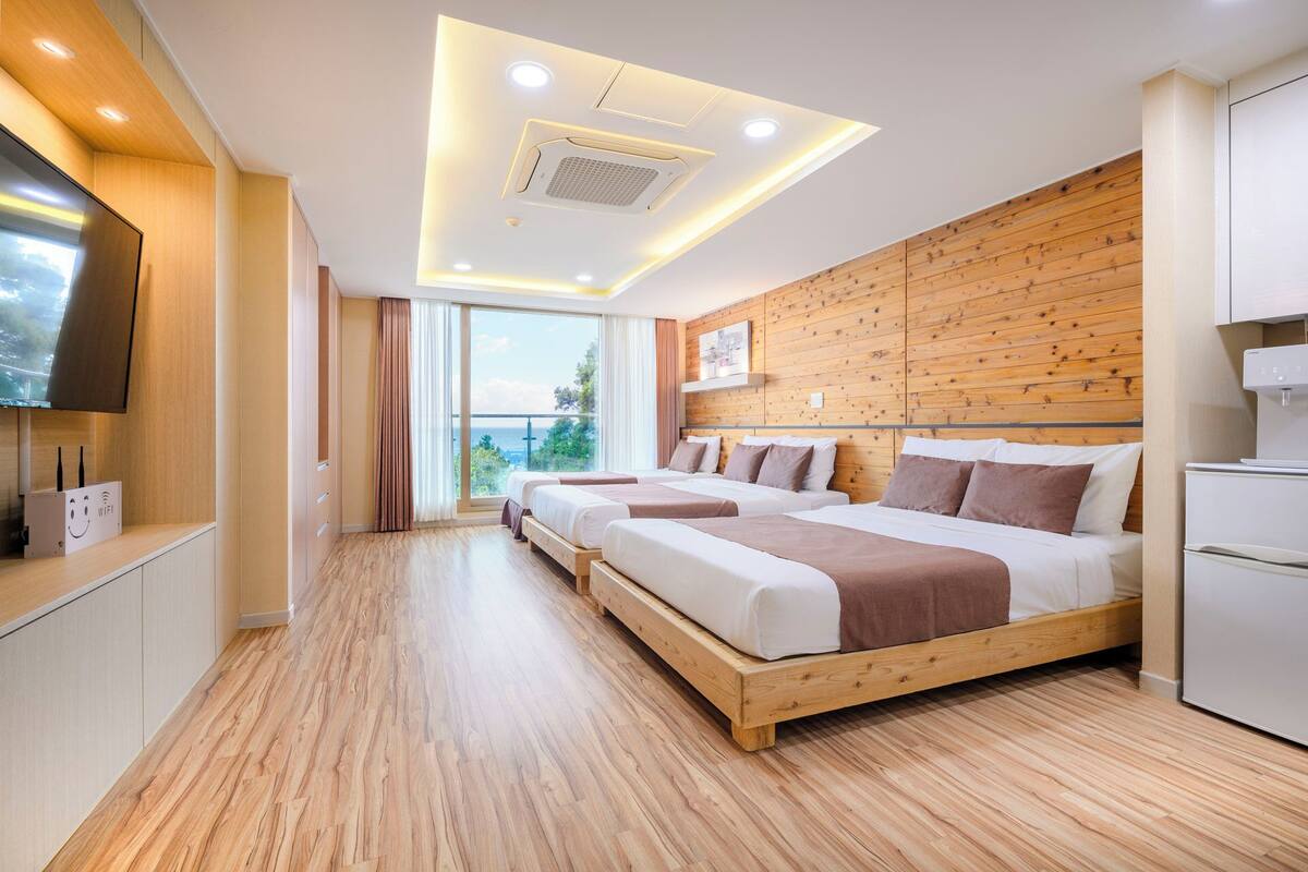 A modern hotel room with a wooden floor, two beds, a TV, and a large window with a view.